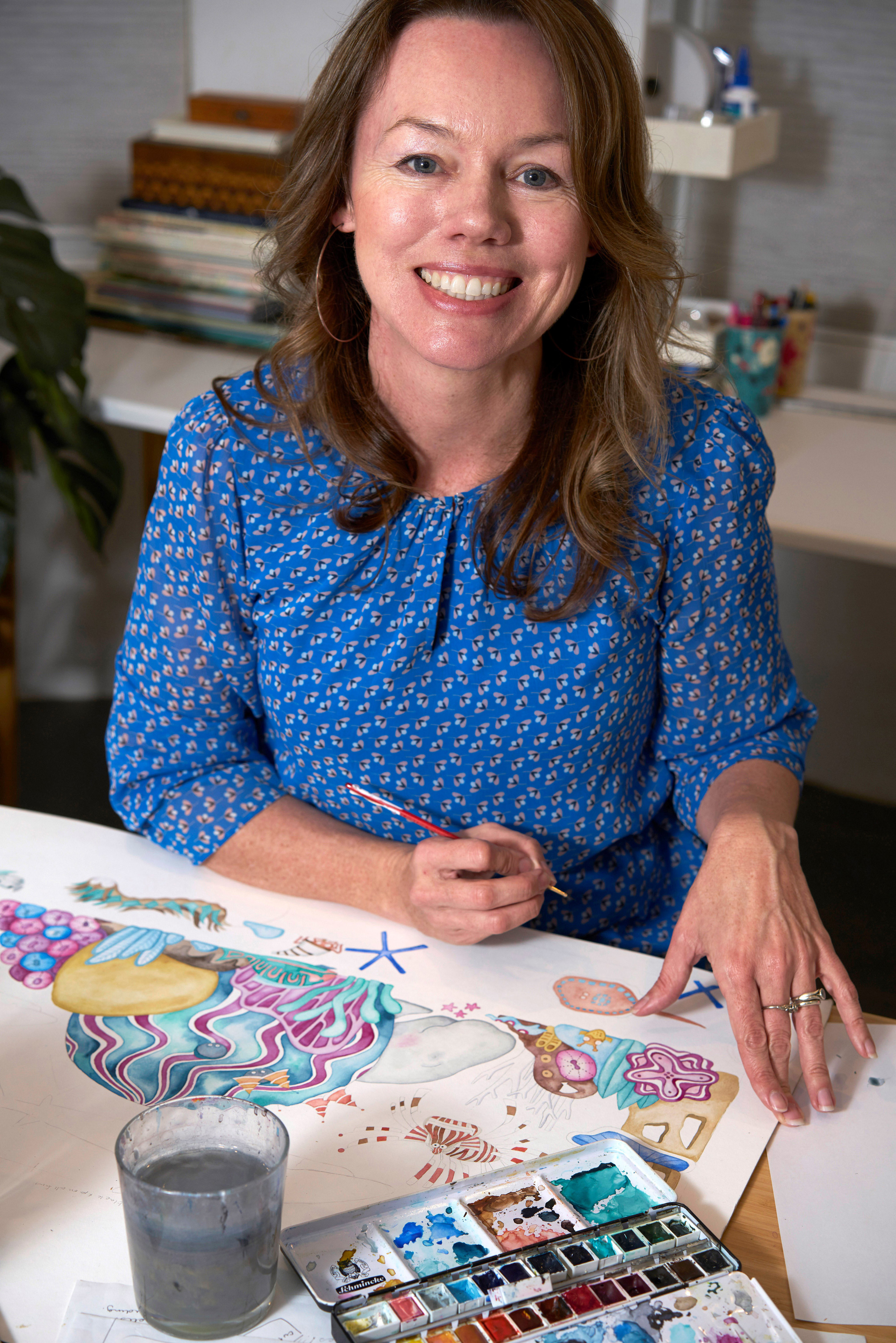 A person with long dark hair, wearing blue, sits at a table with an artwork and art materials