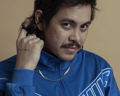 A person with dark hair and a dark moustache wearing a blue zippered top and silver chain