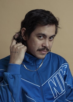 A person with dark hair and a dark moustache wearing a blue zippered top and silver chain