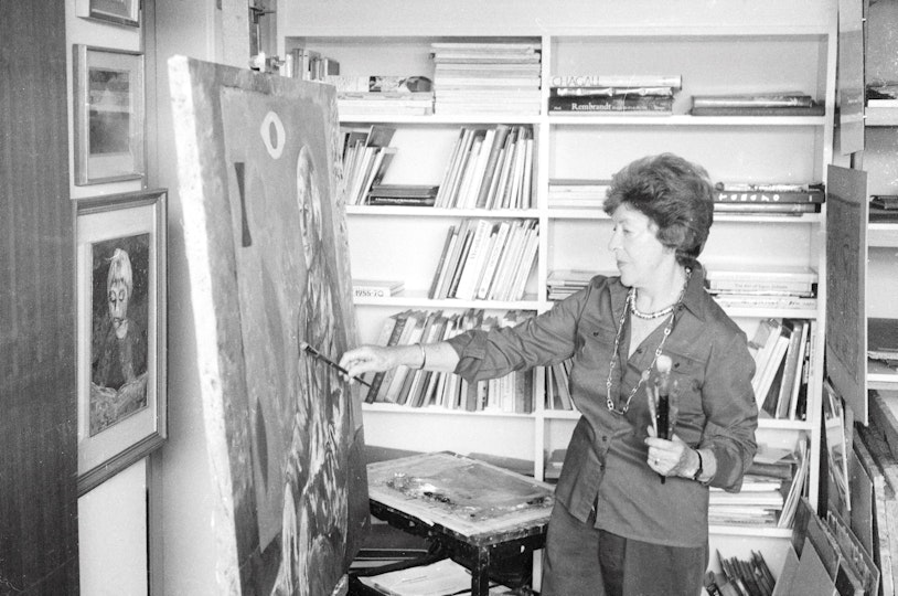 A person holding paintbrushes stands in front of a painting in a room with books on shelves and framed pictures on the wall