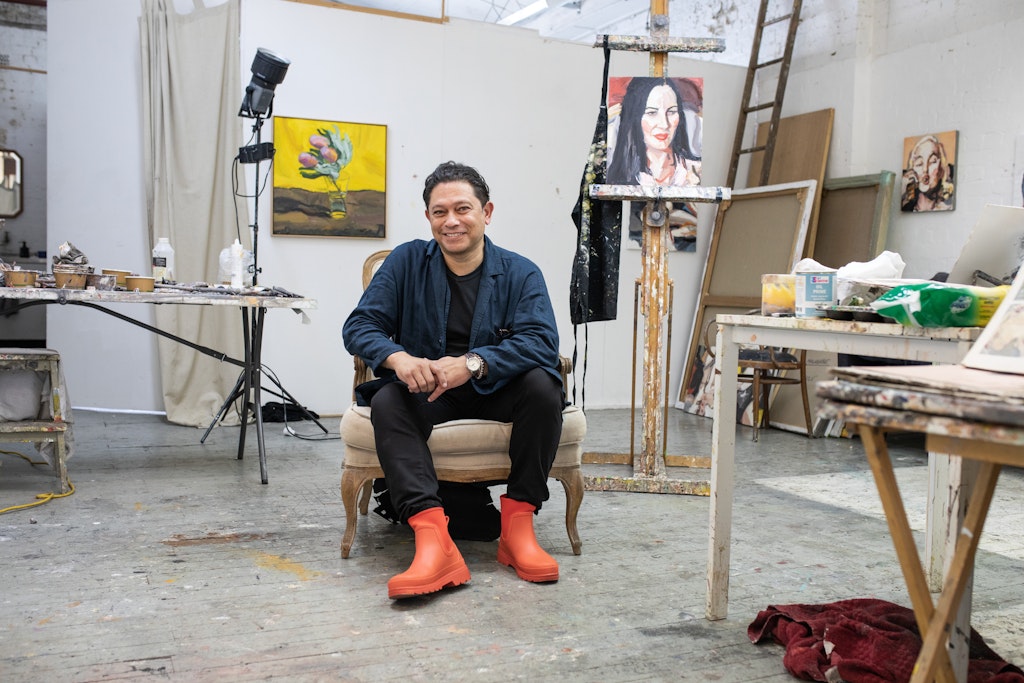 A person sits on an armchair surrounded by paintings and art materials on tables