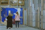 Two people stand in front of a large painting hang with smaller paintings on a wire rack