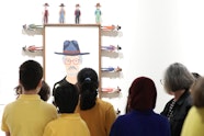 A group of people stand in front of a portrait of a person in a hat and glasses, which is surrounded by small figures wearing hats