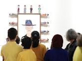 A group of people stand in front of a portrait of a person in a hat and glasses, which is surrounded by small figures wearing hats