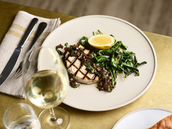 A table setting with knife, fork, napkin, glass of white wine and plate with grilled fish and vegetables