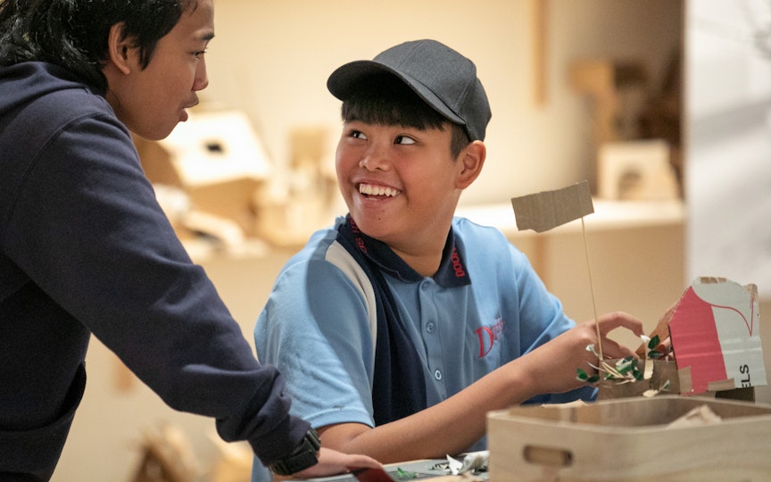 A person sits at a table with a cardboard structure in their hands and looks up smiling at another person leaning towards them