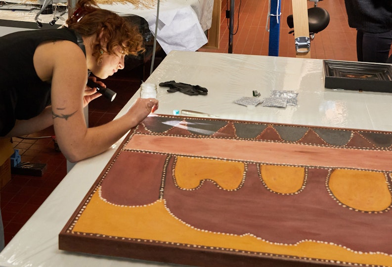 A person leans over a painting lying flat on a workbench