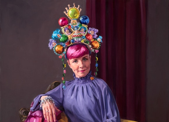 A person with pink hair in a high-necked purple outfit wearing a large colourful headpiece sits in an armchair