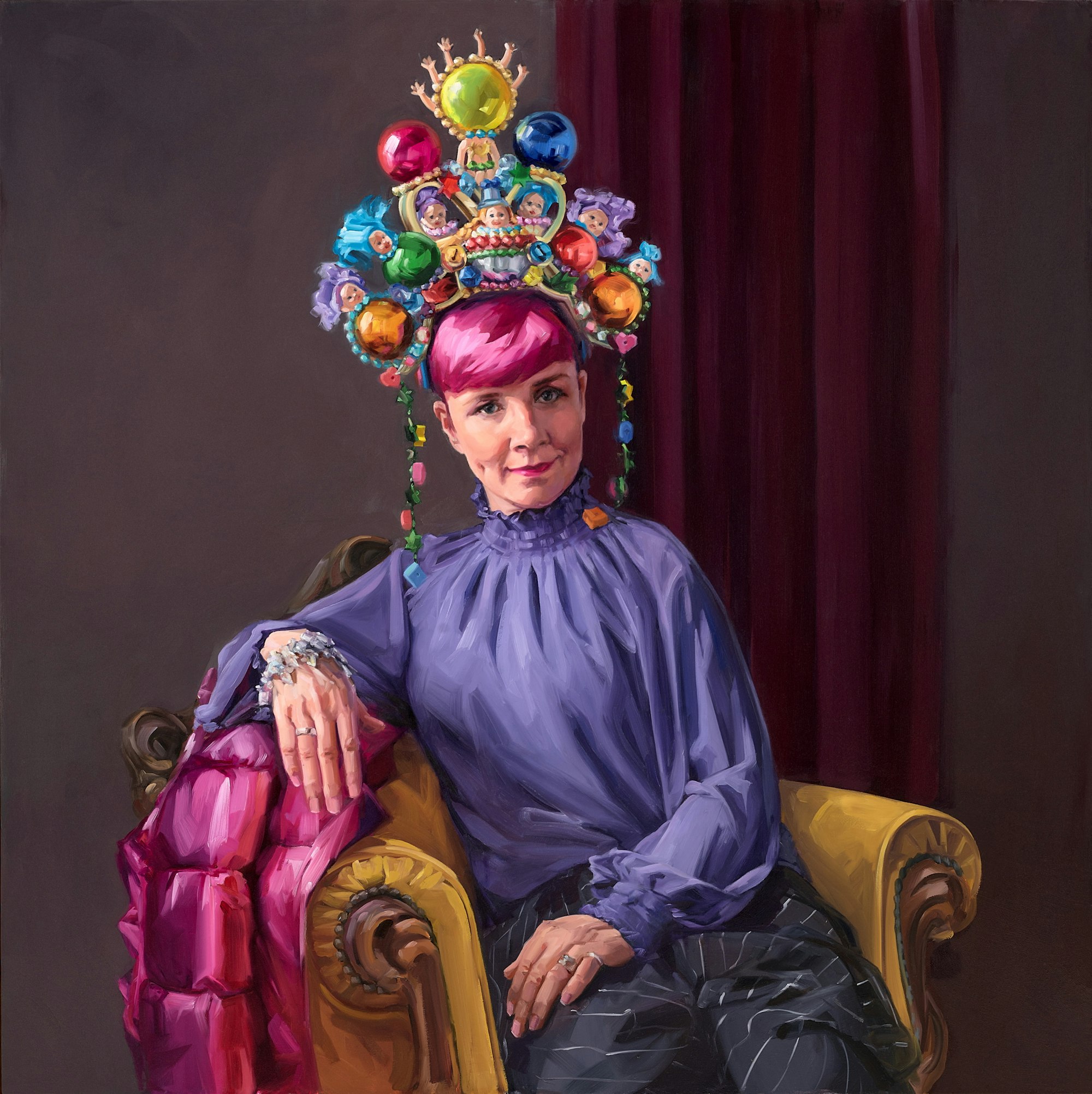 A person with pink hair in a high-necked purple outfit wearing a large colourful headpiece sits in an armchair