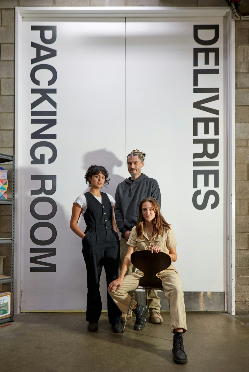 Three people (two standing, one seated) in front of large double doors with the words 'Packing Room Deliveries'