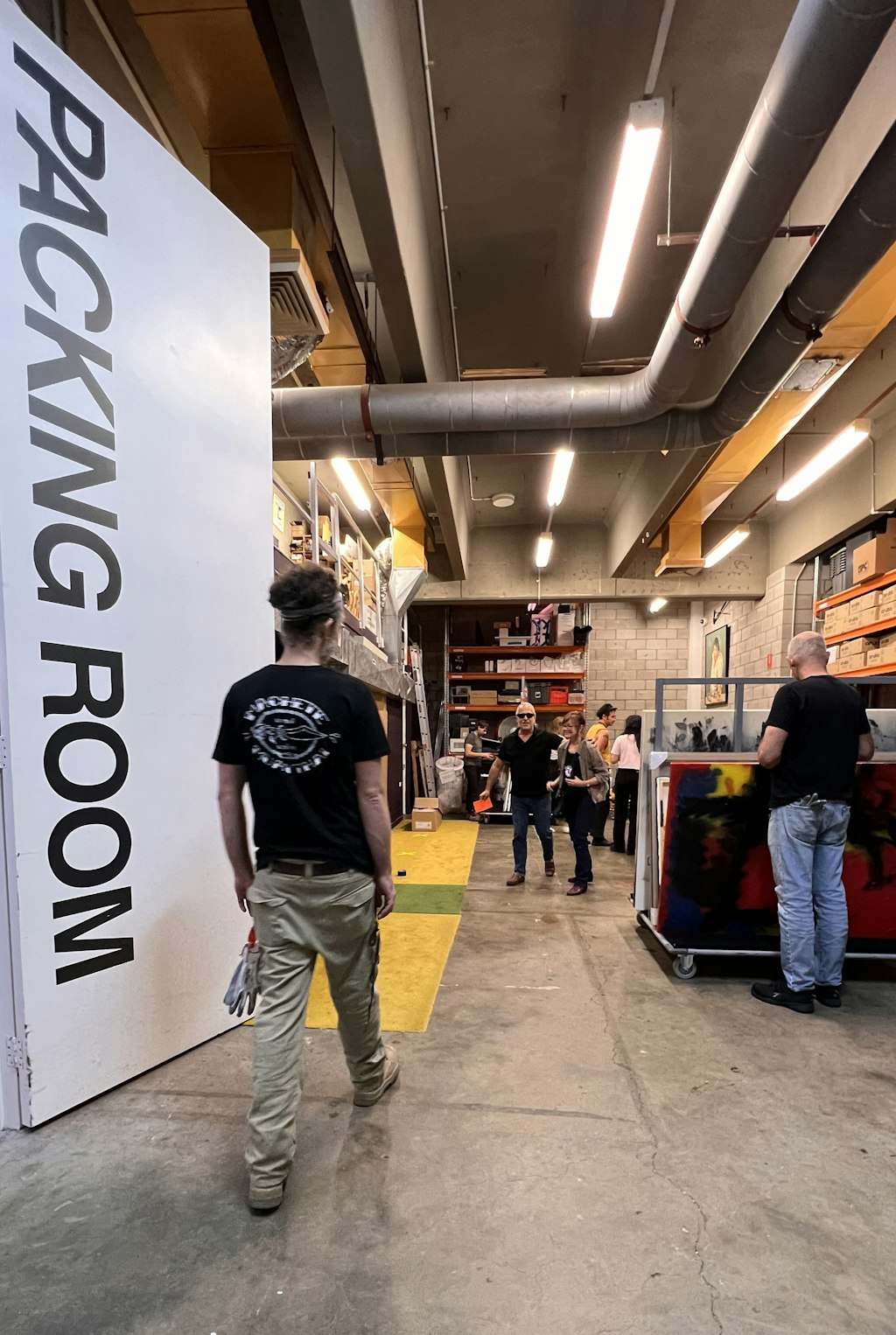 A person walks past a large 'Packing Room' sign into a working space with people, equipment and stored items