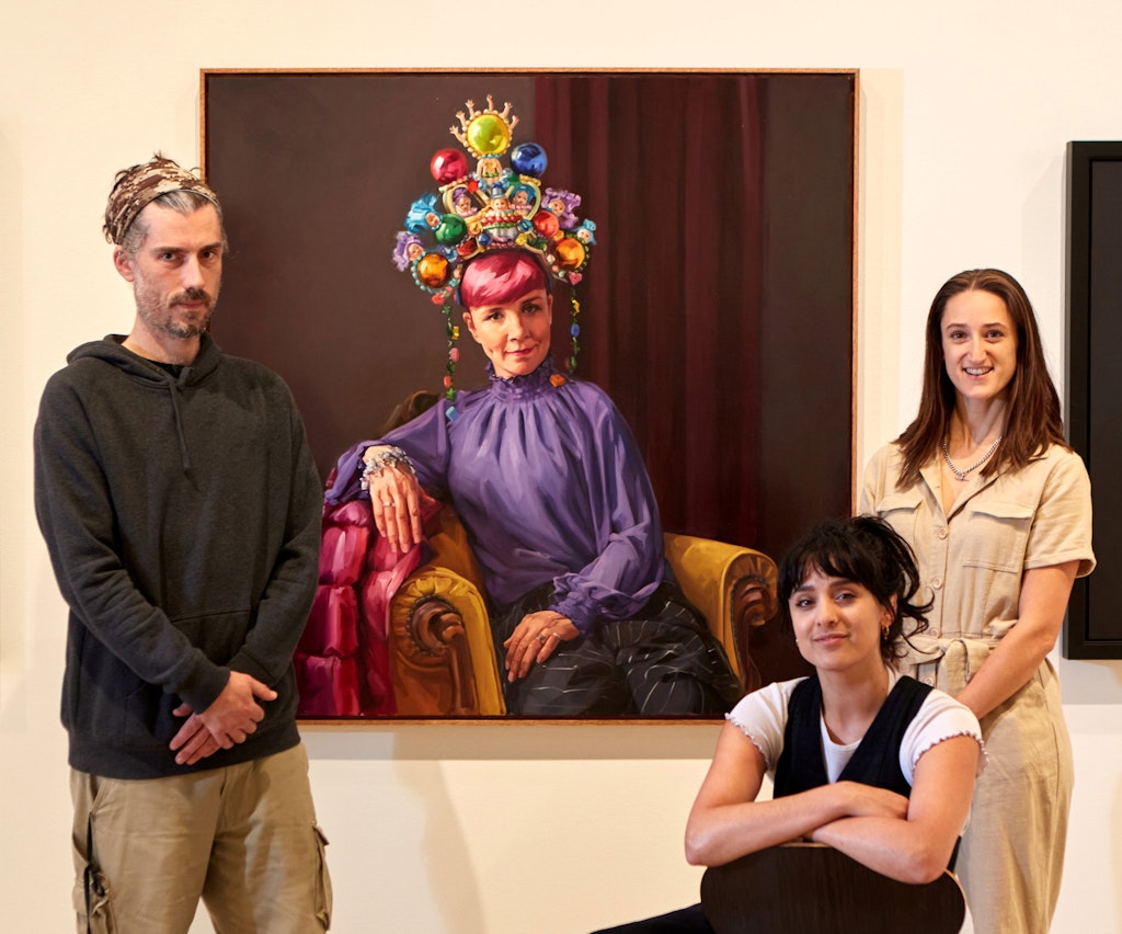 Three people (two standing, one seated) in front of a portrait of a pink-haired person wearing an ornate headpiece