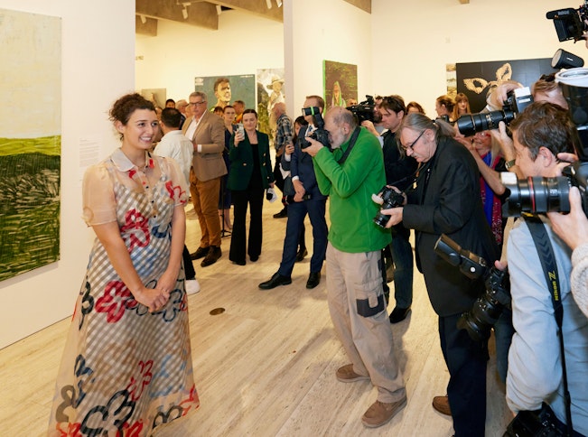 A person stands next to a painting in front of a crowd of people with cameras