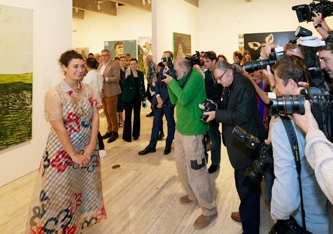 A person stands next to a painting in front of a crowd of people with cameras