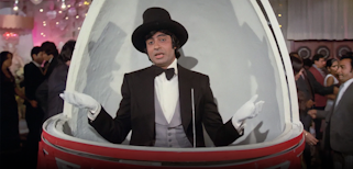A person wearing a hat, gloves, shirt, bow tie, waistcoat and jacket inside a domed structure within a crowded room