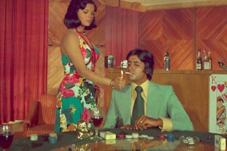 A standing person lights the cigarette of another person seated at a table with gambling chips, a packet of cigarettes and glass in front of them