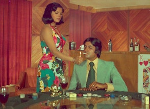 A standing person lights the cigarette of another person seated at a table with gambling chips, a packet of cigarettes and glass in front of them