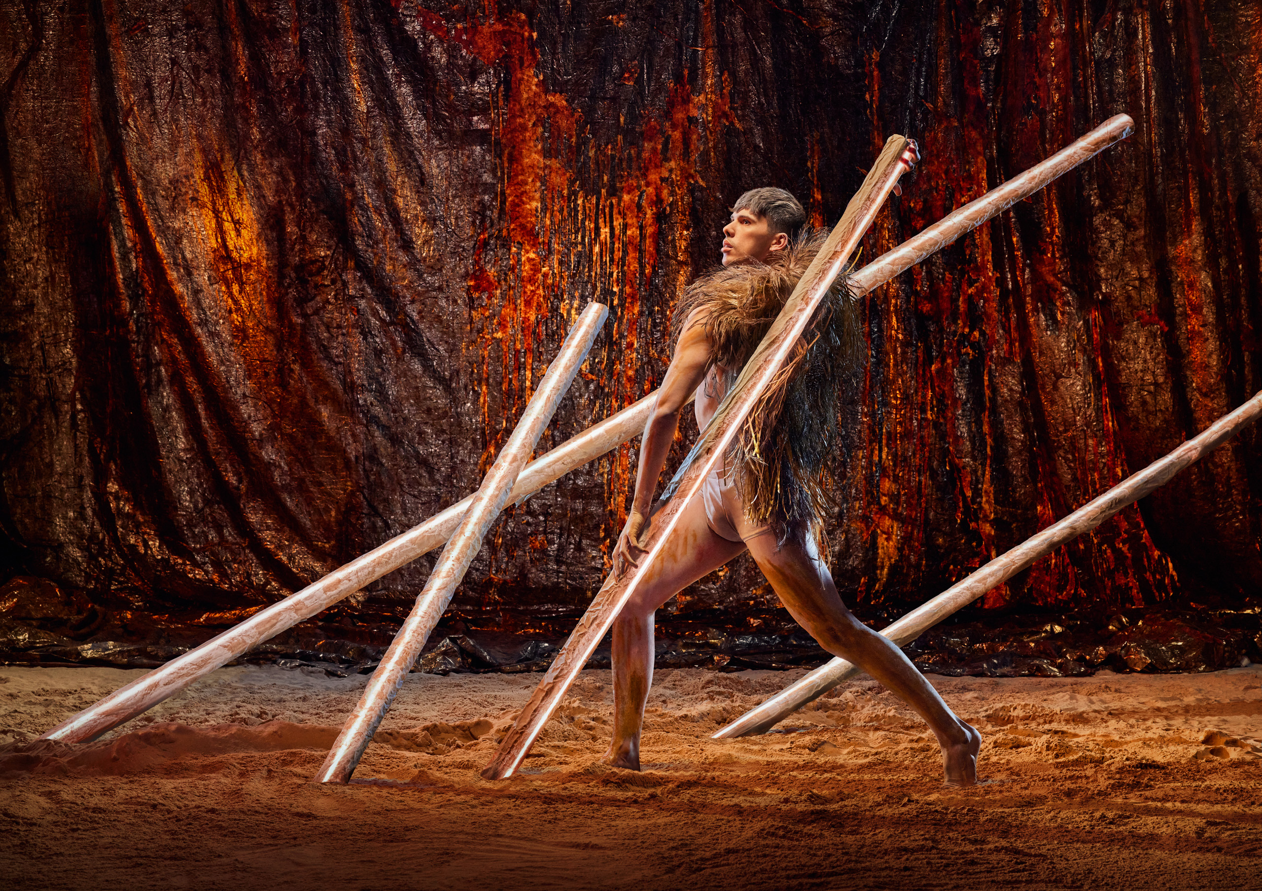 In front of a black and orange curtain, a person wearing a feather cloak lunges between four poles that jut at angles from sand