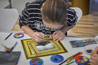 A person sits leaning over an art project in progress, surrounded by paints and other art materials