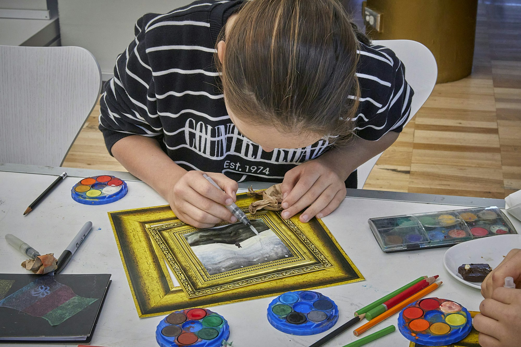 A person sits leaning over an art project in progress, surrounded by paints and other art materials