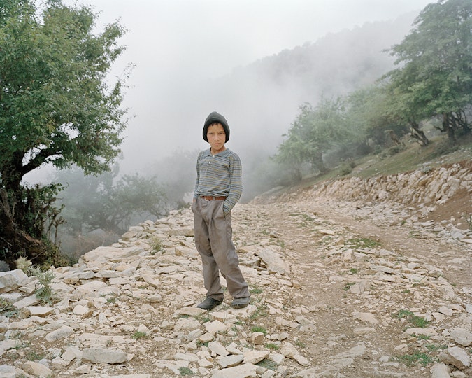 A young person, wearing a hat, stands with their hands in their trouser pockets on a rocky road on a hillside