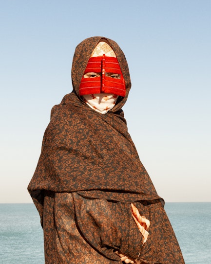A person swathed in fabric over their head and body wears a red face covering revealing only their eyes
