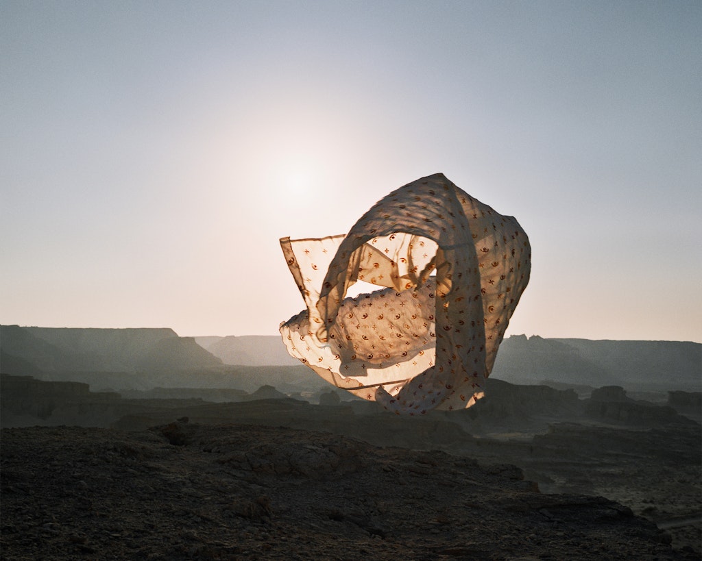 A figure within a long billowing piece of fabric within a desert landscape in half light