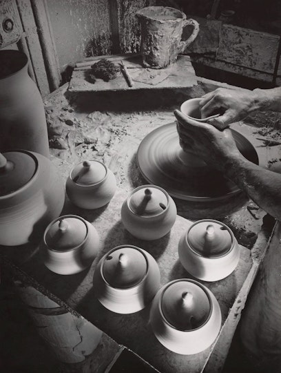 Two hands at a potter's wheel with formed ceramic vessels in the foreground and art-making materials in the background