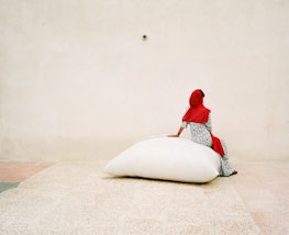 A person wearing a bright red headscarf sits on a large white bag on a pale-coloured ground in front of a pale wall