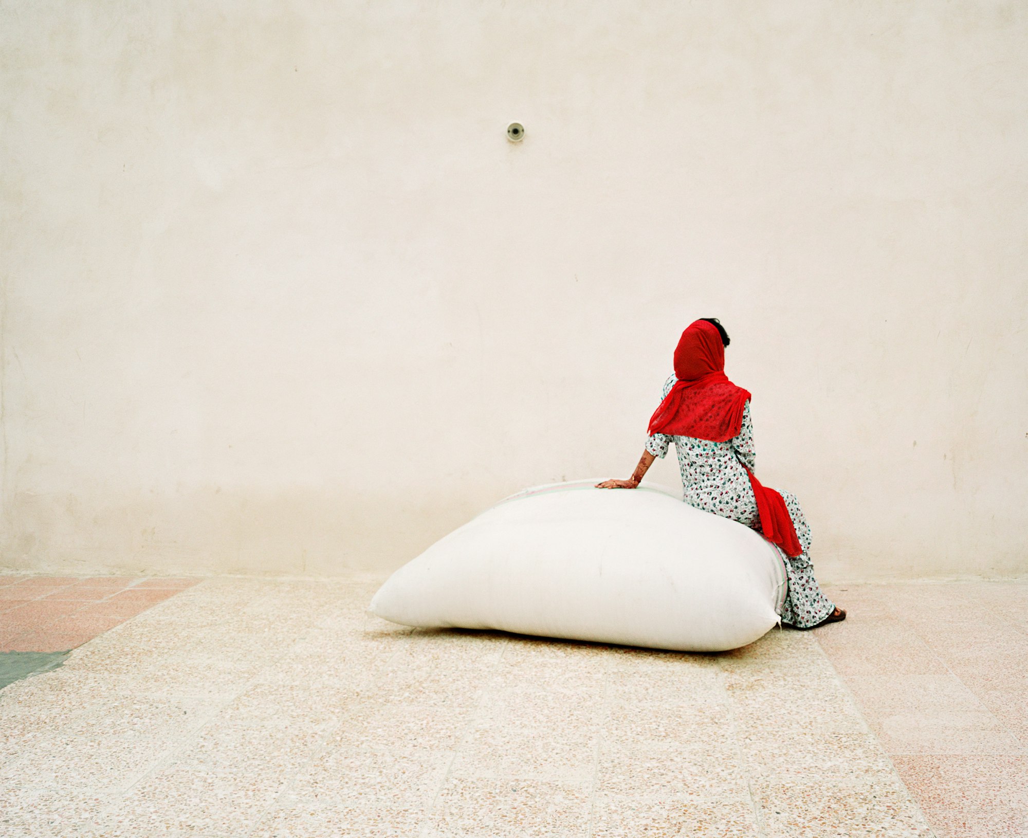 A person wearing a bright red headscarf sits on a large white bag on a pale-coloured ground in front of a pale wall