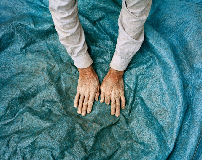 A pair of hands, missing one index finger, of someone in a pale long-sleeved garment on a wrinkled blue ground cover