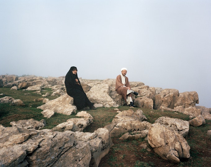 Two people in head coverings and long robes sit on a rocky outcrop under a grey misty sky