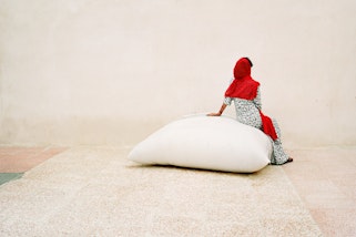 A person wearing a bright red headscarf sits on a large white bag on a pale-coloured ground in front of a pale wall