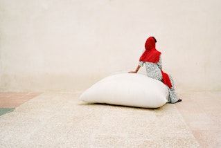 A person wearing a bright red headscarf sits on a large white bag on a pale-coloured ground in front of a pale wall