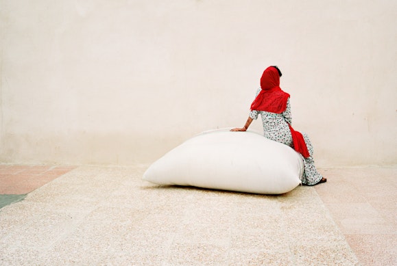 A person wearing a bright red headscarf sits on a large white bag on a pale-coloured ground in front of a pale wall