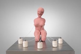 A plinth with a kneeling pink figure which has five spools of thread attached to its nipples