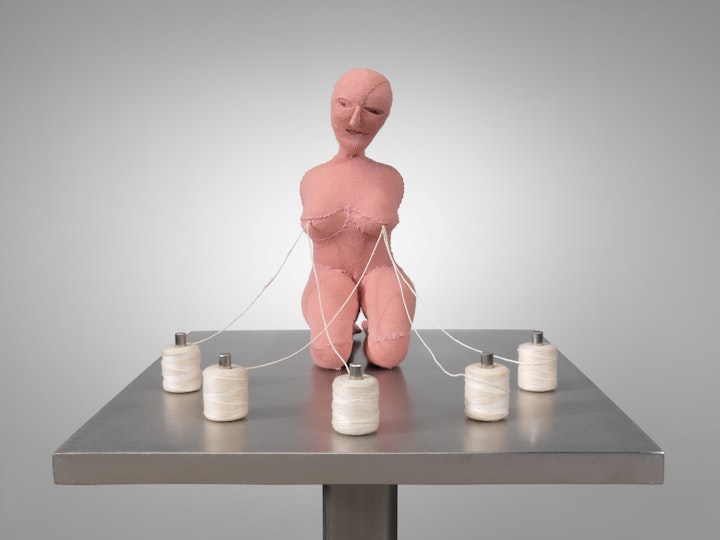 A plinth with a kneeling pink figure which has five spools of thread attached to its nipples