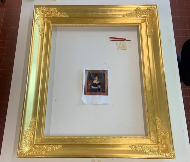 A gold frame lies on a cardboard surface with a small paper reproduction of a framed portrait