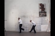 A film still of two older people walking in front of a studio wall.