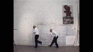 A film still of two older people walking in front of a studio wall.