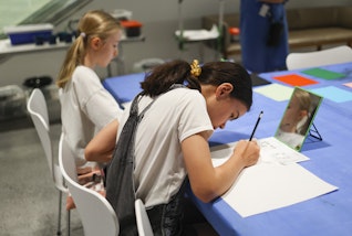 Two people seated at a table. One is holding a pencil to a piece of paper with a mirror propped in front of them