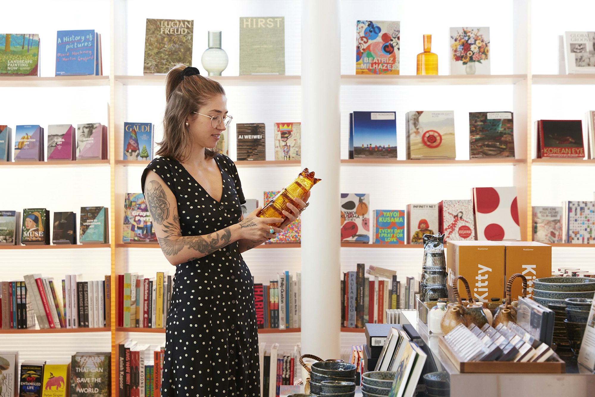 A person stands holding an object in front of bookshelves and a display of objects