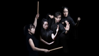 Four people dressed in black; one holding drum sticks and the others with their hands in martial arts poses