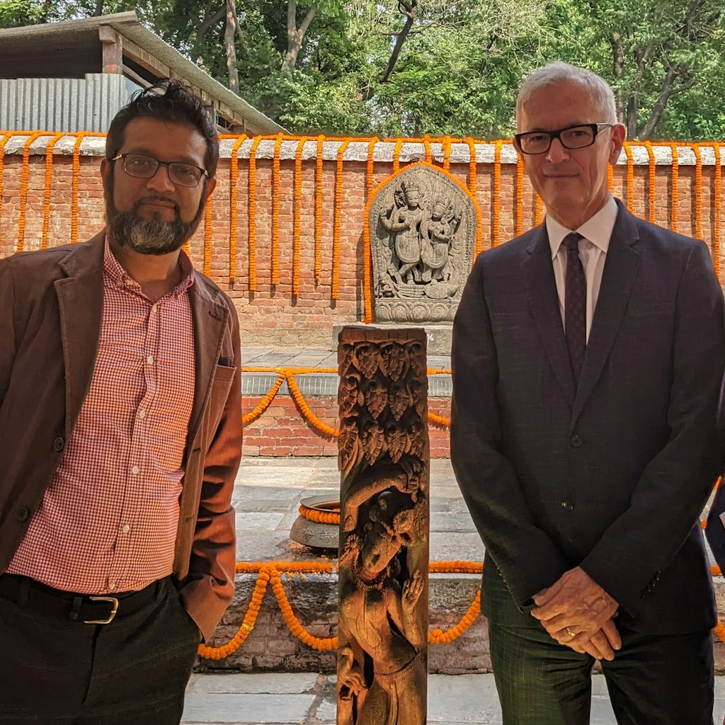 Two people stand either side of a carved wooden column. Another sculpture is in the distance behind them