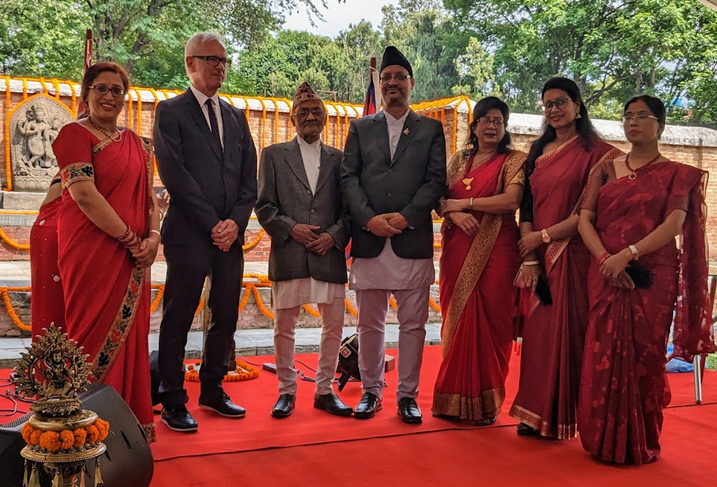 Seven people standing side by side on red carpet. Four are dressed in red saris; the other three are in jackets and pants.