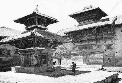 A black-and-white photo of a narrow, three-story building, flanked by other buildings, with children and animals in front of it.