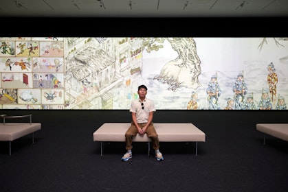 A person sits on a bench in front of a large screen showing an illustrated artwork