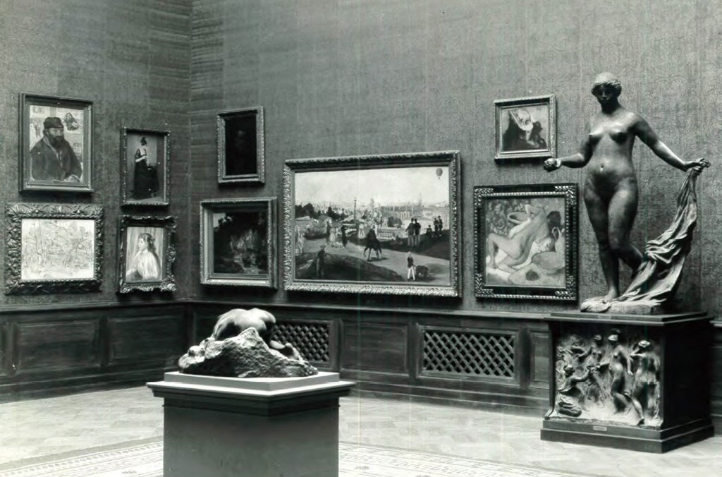 The corner of a gallery room with framed paintings on the wall and two sculptures on pedestals