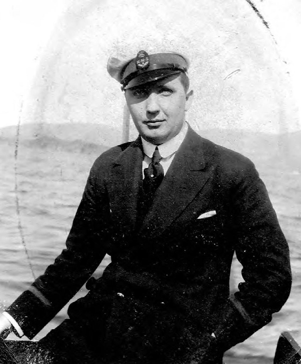 A person in a jacket, shirt, tie and sailors cap with choppy water in the background