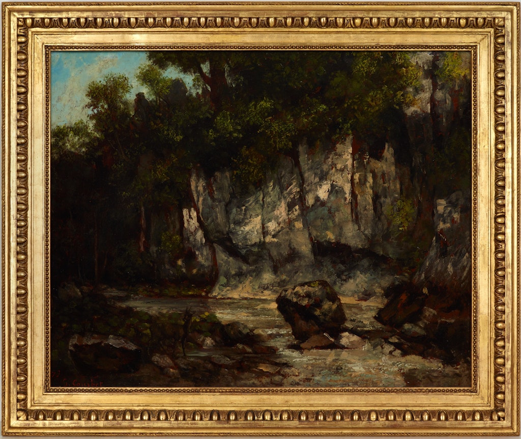 A gold-framed painting of a rocky precipice in a wooded landscape with a boulder in the foreground